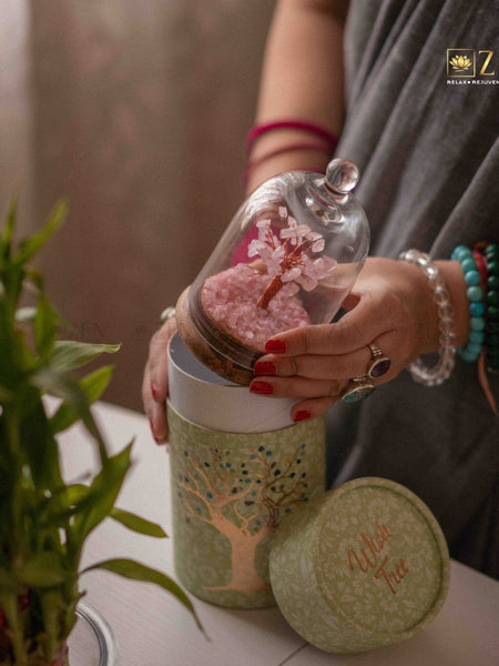 Person holding a decorative jar with a pink tree inside, next to a plant on a table.