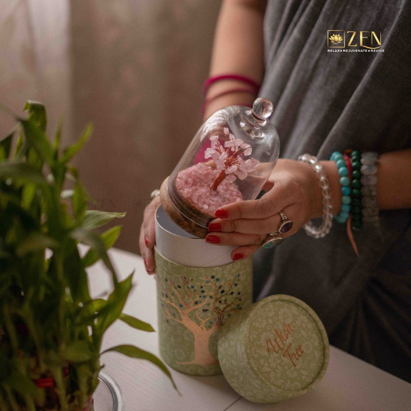 Person holding a decorative jar with a pink tree inside, next to a plant on a table.