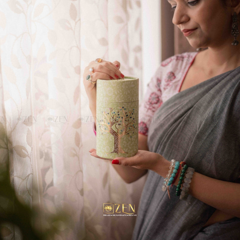 Woman holding a cylindrical container with a tree design, wearing a gray saree and colorful bracelets.
