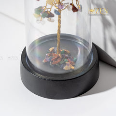 Decorative item with multicolored stones and a gold branch inside a glass dome on a white background.
