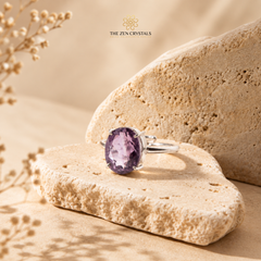 A silver ring with an oval purple amethyst crystal setting, displayed on a textured beige stone against a warm sandy background. The brand logo 'THE ZEN CRYSTALS' is visible at the top center.