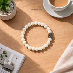 mother of pearl bracelet with Buddha charm on a wooden table with a cup of coffee and plant in the background.