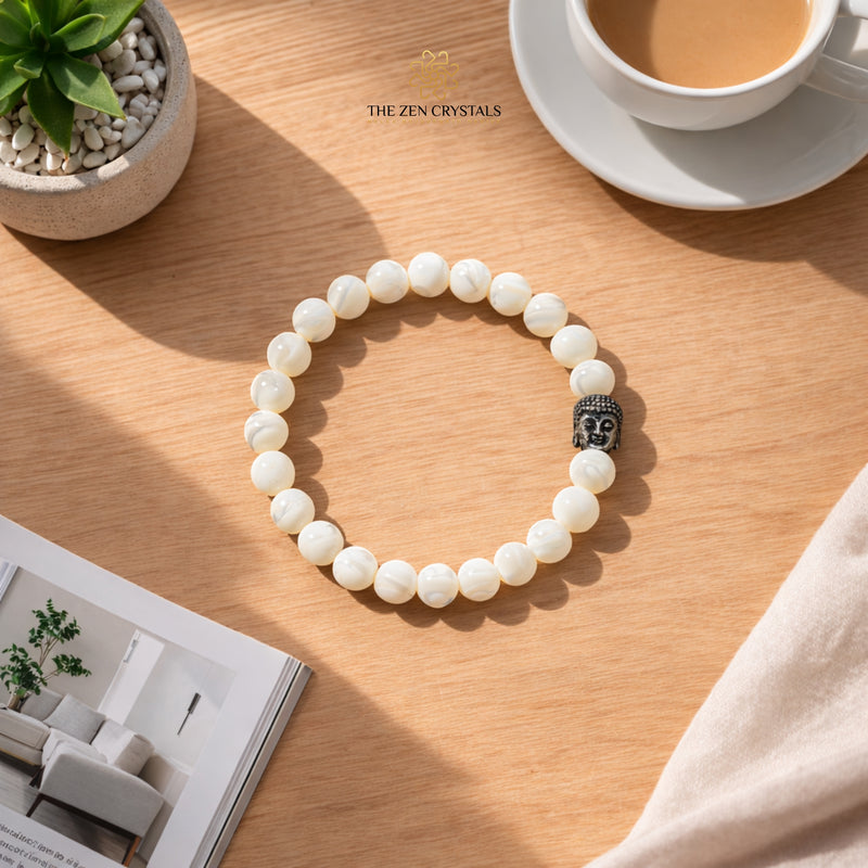 mother of pearl bracelet with Buddha charm on a wooden table with a cup of coffee and plant in the background.