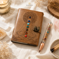 A brown leather journal with an embossed meditating figure and chakra design, shown on a draped fabric surface with a matching chakra-themed pen, crystals, dried flowers, and a candle. The brand name 'THE ZEN CRYSTALS' is visible at the top.