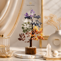 A multicolor 7 chakra crystal tree of life in a wooden base, displayed on a marble tray on a marble table with decorative dried flowers and crystals in a warm, elegant setting. The brand logo 'THE ZEN CRYSTALS' is visible at the top.