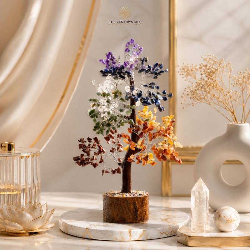 A multicolor 7 chakra crystal tree of life in a wooden base, displayed on a marble tray on a marble table with decorative dried flowers and crystals in a warm, elegant setting. The brand logo 'THE ZEN CRYSTALS' is visible at the top.