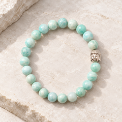 An amazonite bracelet made of round light green and white marbled beads with a silver Buddha head charm, displayed on a light beige stone surface.