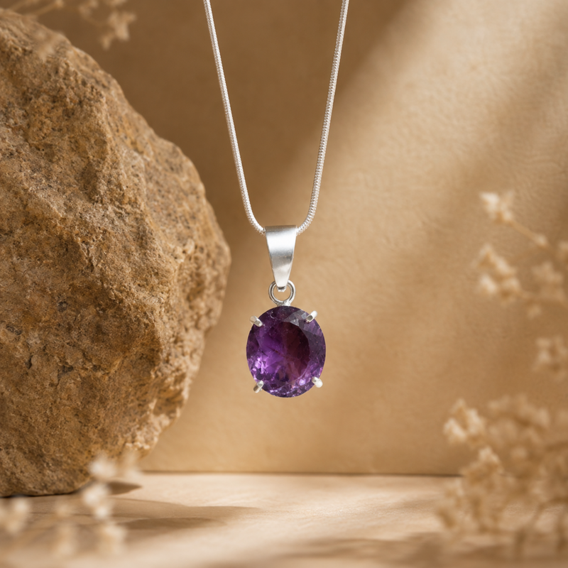 A Amethyst pendant necklace with an oval purple gemstone, displayed against a warm beige background with a textured rock and dried floral elements.