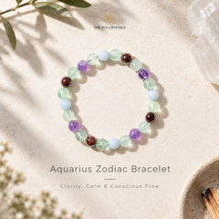 A beaded stretch bracelet for the Aquarius zodiac, featuring translucent light green, lavender purple, pale blue, and dark brown gemstone beads. The bracelet is displayed on a textured beige surface with soft shadows, alongside decorative elements like a crystal cluster, dried flowers, and a bowl of matches.