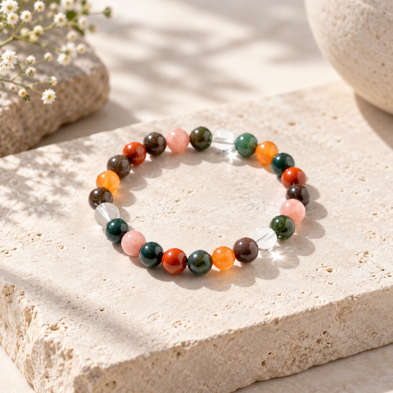 An aries zodiac bracelet made of polished multicolored gemstone-like beads, including orange, green, pink, red, and dark brown, with a few clear faceted beads, displayed on a textured beige stone surface.