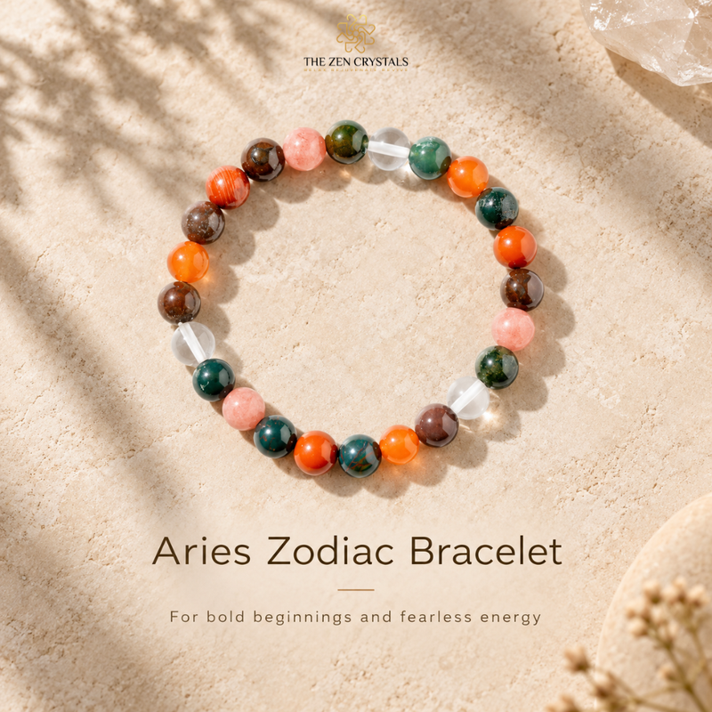 A beaded stretch bracelet labeled 'Aries Zodiac Bracelet' by The Zen Crystals, featuring marbled orange, green, brown, and pink gemstone beads with clear crystal accents, displayed on a sandy beige surface with soft leaf shadows.