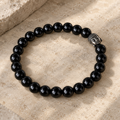 A black obsidian bracelet with a polished silver Buddha head charm, displayed on a textured beige stone surface.