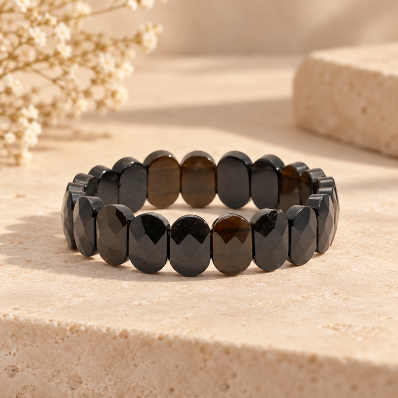 Faceted Black Obsidian bracelet arranged in a circle on a light beige stone surface with dried flowers in the background.