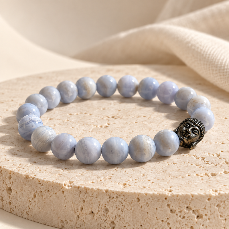 A blue lace agate bracelet made of light blue-gray round beads with a dark metallic Buddha head charm, displayed on a beige stone slab with a soft cream fabric background.
