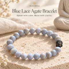 A beaded bracelet made of pale blue-gray agate-like round beads with a small black Buddha head charm, displayed on a textured beige stone surface with soft fabric and dried flowers in the background. Text at the top reads 'Blue Lace Agate Bracelet' and 'The Zen Crystals'.