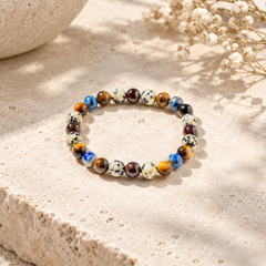 A beaded stretch bracelet made of multicolored polished gemstone beads, resting on a textured beige stone surface with dried flowers in the background.