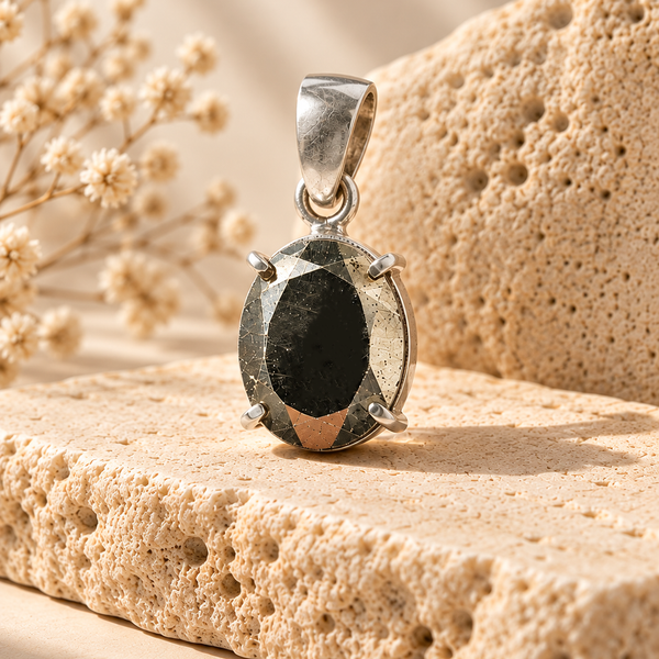 A faceted Pyrite pendant with a polished silver bail, set in a four-prong setting, displayed on a textured beige stone with dried flowers in the background.