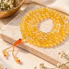 citrine jaap mala on a light stone slab with small white flowers and a gold bowl in the background.