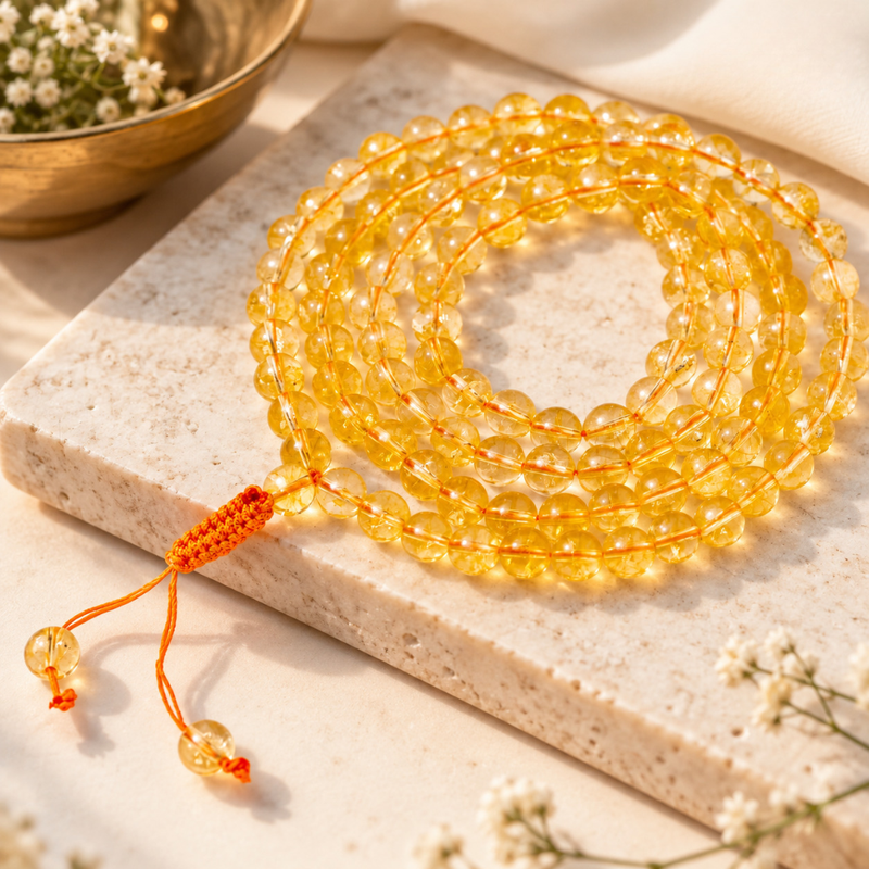 citrine jaap mala on a light stone slab with small white flowers and a gold bowl in the background.
