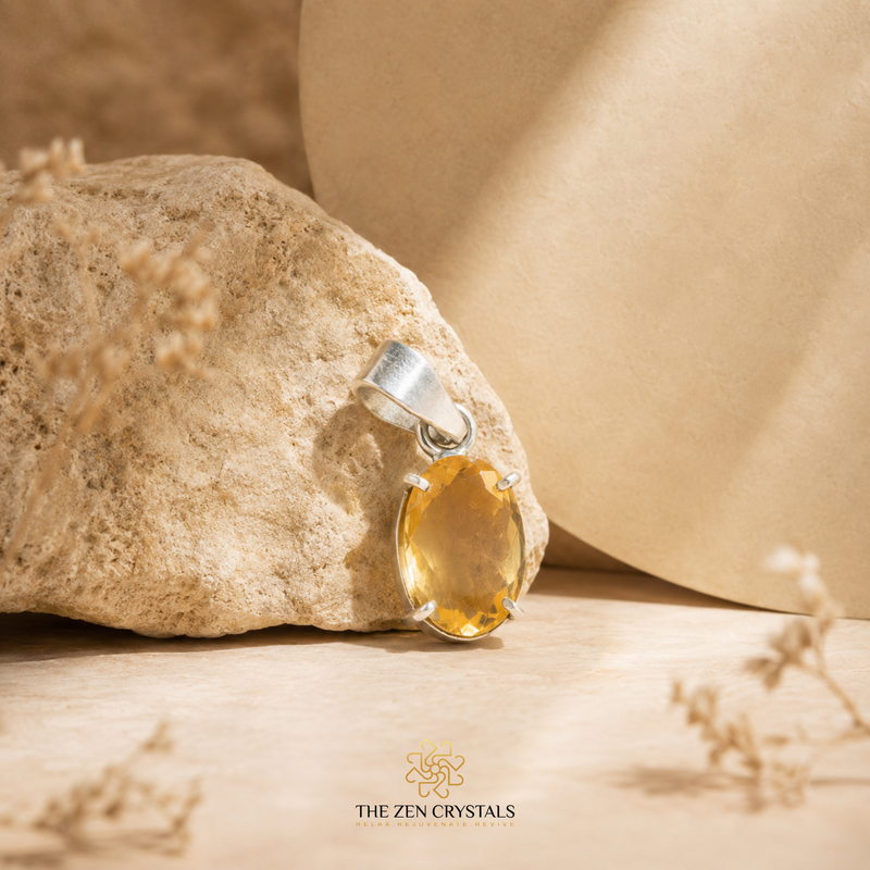 A faceted citrine pendant resting on a textured beige rock, with a soft beige background and dried floral elements. The brand logo 'THE ZEN CRYSTALS' is visible at the bottom.