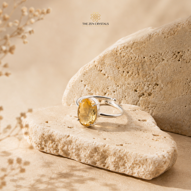 A silver ring with a faceted yellow crystal gemstone resting on a textured beige stone, with a second stone behind it and the brand name 'THE ZEN CRYSTALS' printed above.