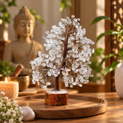 A crystal tree of life decorative sculpture made of clear faceted stones on a dark brown wire branch, displayed on a round wooden tray in a warm, Zen-style room with a Buddha statue and plants in the background.