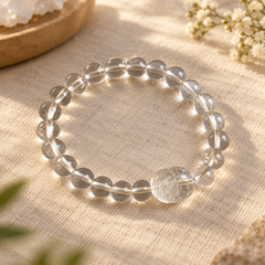 A clear crystal beaded bracelet with a large polished round bead, displayed on a textured beige linen fabric with soft natural shadows.