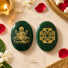 Two polished dark green crystal stones with gold engravings, placed on a light marble surface with scattered rose petals, white flowers, a lit candle, and ornate gold-toned decorative objects around them. The left stone shows a seated multi-armed deity motif, and the right stone shows a geometric mandala design.