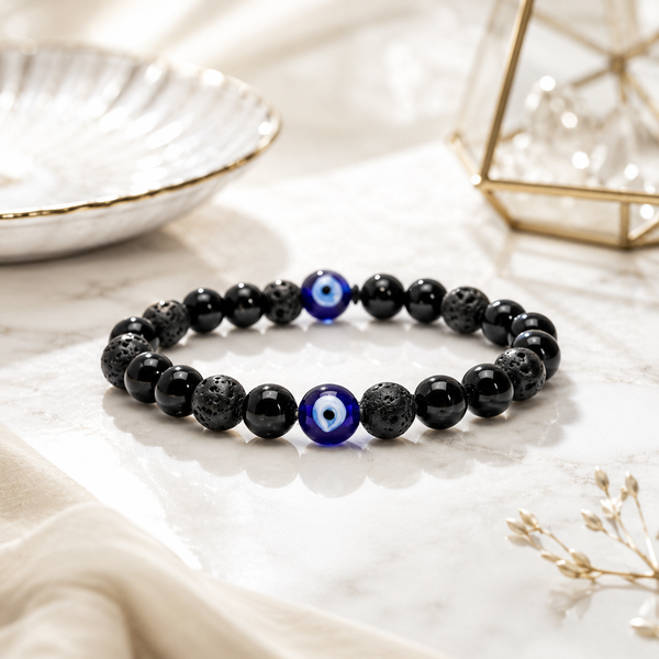 Evil Eye bracelet featuring black lava stone beads and a central blue evil eye charm, displayed on a white marble surface with soft-focus decorative props.