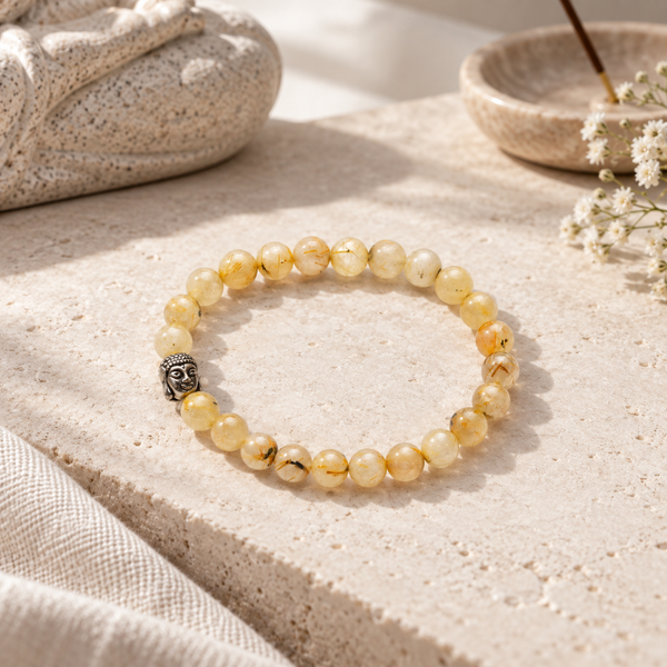 A Golden Rutile bracelet made of translucent pale yellow beads with natural inclusions, featuring a small silver-toned Buddha head charm, displayed on a textured beige stone surface.