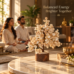 A decorative flowering tree sculpture made of cream-colored petals on a rustic brown base, placed on a marble table in a warm, sunlit living room. Two people are seated in the background in soft focus, and text in the upper right reads 'Balanced Energy Brighter Together.