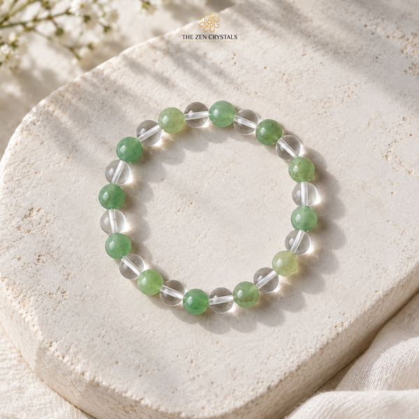 A beaded stretch bracelet by The Zen Crystals, featuring alternating translucent clear crystal beads and round green jade-like beads, displayed on a textured beige stone slab.