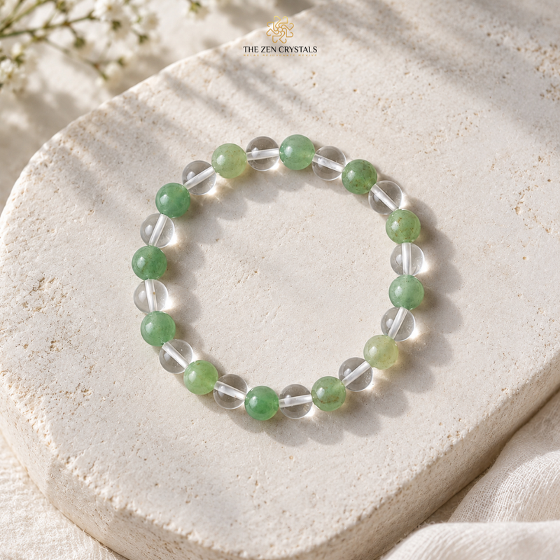 A beaded stretch bracelet by The Zen Crystals, featuring alternating translucent clear crystal beads and round green jade-like beads, displayed on a textured beige stone slab.