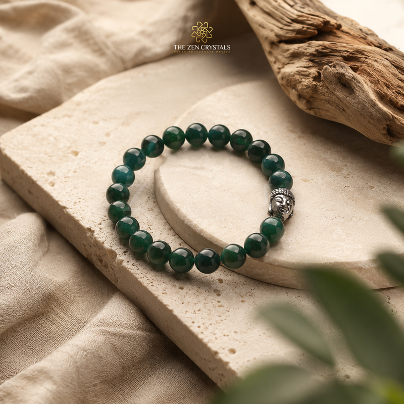 Natural Jade bracelet made of dark green faceted gemstone beads with a silver Buddha head charm, displayed on a textured beige stone slab with a piece of driftwood and soft fabric in the background. The brand logo 'THE ZEN CRYSTALS' is visible at the top.