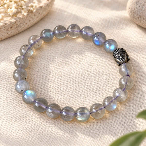A Labradorite bracelet featuring round gray labradorite beads with a blue iridescent sheen, a purple cord, and a dark bronze Buddha head charm, displayed on a textured beige linen fabric.