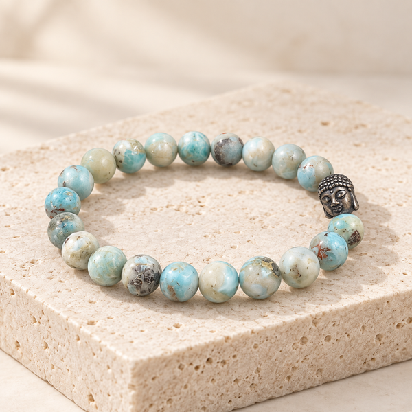 A beaded stretch larimar bracelet made of round turquoise-blue and cream marbled beads, with a small dark metallic Buddha head charm, displayed on a beige stone block.