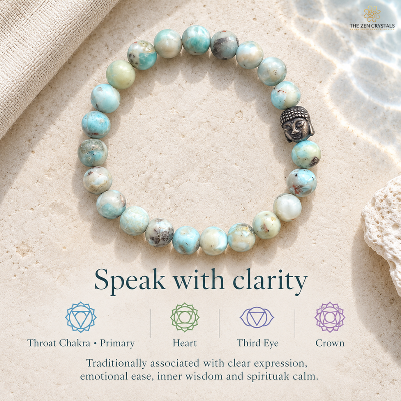 A beaded stretch larimar bracelet made of light blue and white marbled beads with a small black Buddha head bead, displayed on a sandy surface with water ripples and text explaining chakra associations.