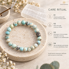 A beaded bracelet made of light blue and green marbled stones with a small black Buddha bead, displayed on a beige stone plate. The image includes a 'Care Ritual' guide on the right side with text and icons, and decorative props like a crystal, incense, and dried flowers.
