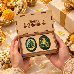 A pair of oval-shaped green and gold Diwali-themed earrings held in an open wooden gift box with 'Happy Diwali' printed on the inside lid. Two hands are visible holding the box, surrounded by festive decorations including lit candles, flowers, and a gold ribboned gift box in the background.