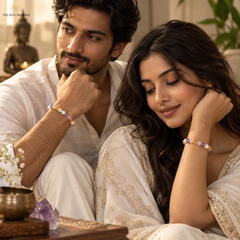 A man and a woman in white traditional clothing sit closely together, each wearing a beaded bracelet with pastel purple, yellow, and white beads. The scene is softly lit with warm tones and includes decorative objects like a brass bowl, crystal, and Buddha statue in the background.