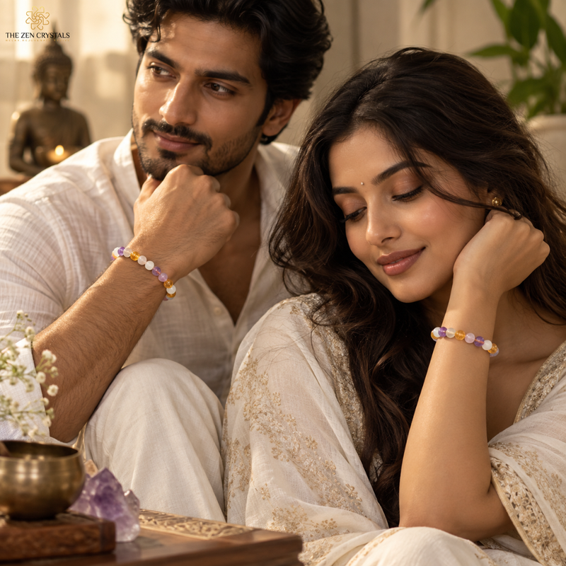 A man and a woman in white traditional clothing sit closely together, each wearing a beaded bracelet with pastel purple, yellow, and white beads. The scene is softly lit with warm tones and includes decorative objects like a brass bowl, crystal, and Buddha statue in the background.