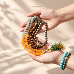 A pair of hands holding several strands of beaded prayer ropes and matching bracelets from The Zen Crystals. The beads are made of various natural stones in shades of gray, green, brown, orange, and turquoise, with one tassel featuring green and orange threads.