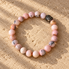 A pink opal bracelet made of round pink and white marbled beads with a small black metal Buddha head charm, displayed on a sandy surface.