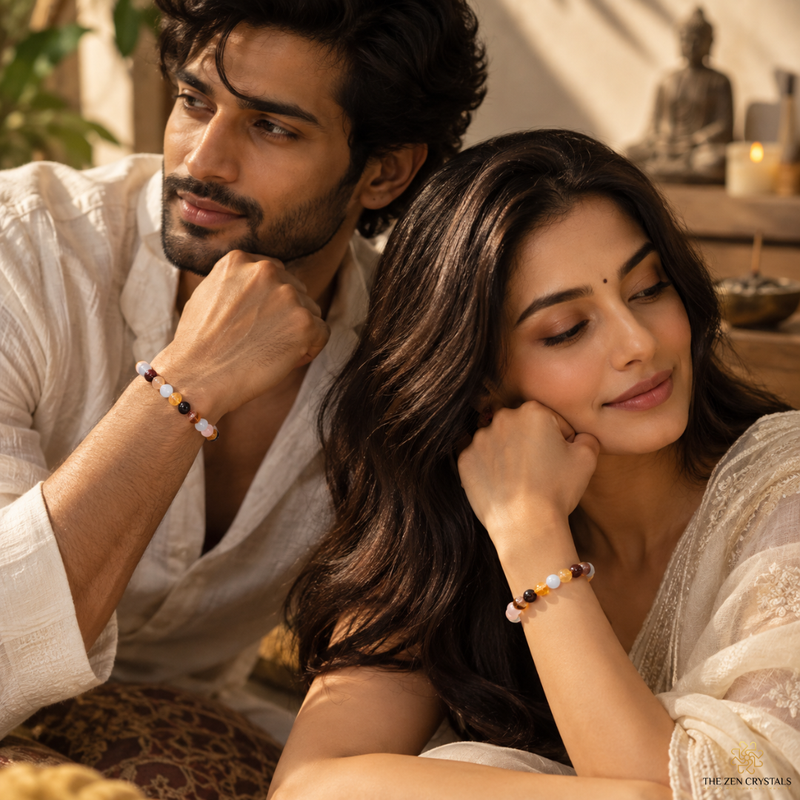 A man and a woman in white traditional clothing sit closely together, each wearing a small beaded bracelet. The scene is warmly lit with a soft-focus background containing a Buddha statue and candles.
