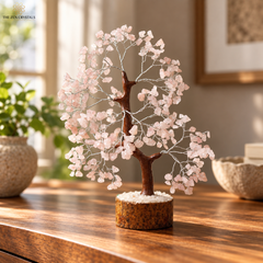 A rose quartz crystal tree decorative piece with a brown wire trunk and branches adorned with pale pink rose quartz leaves, mounted on a round textured base, displayed on a wooden table in a warm, softly blurred interior setting.