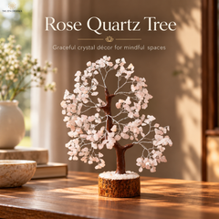 A rose quartz crystal tree decorative piece displayed on a wooden table in a warm, sunlit room. The tree features a dark brown wire branch structure adorned with numerous pale pink rose quartz leaves, mounted on a round cork base with white stones on top.