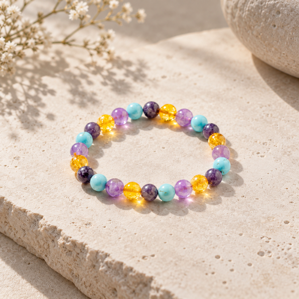 A beaded stretch bracelet with alternating translucent purple, light blue, yellow-orange, and opaque lavender beads, displayed on a textured beige stone surface.