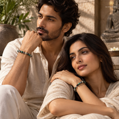 A man and a woman in matching cream-colored linen clothing sit closely together in a warm, sunlit indoor setting. Each wears a beaded bracelet with dark green and metallic beads.