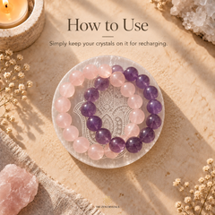 A pair of beaded crystal bracelets, one pink and one purple, displayed on a clear glass plate over a white Hamsa hand. The scene is styled with a candle, dried flowers, and raw crystal rocks on a textured beige surface, with the text 'How to Use' and 'Simply keep your crystals on them for recharging.