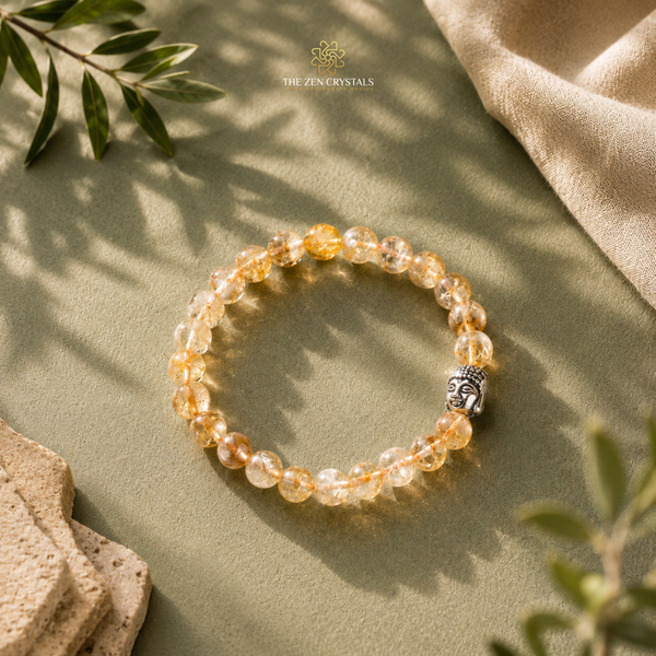 A Unheated Citrine 8mm bracelet made of clear faceted crystals with warm golden-orange tones and a small silver Buddha head charm, styled on a sage green surface with leaves and fabric.